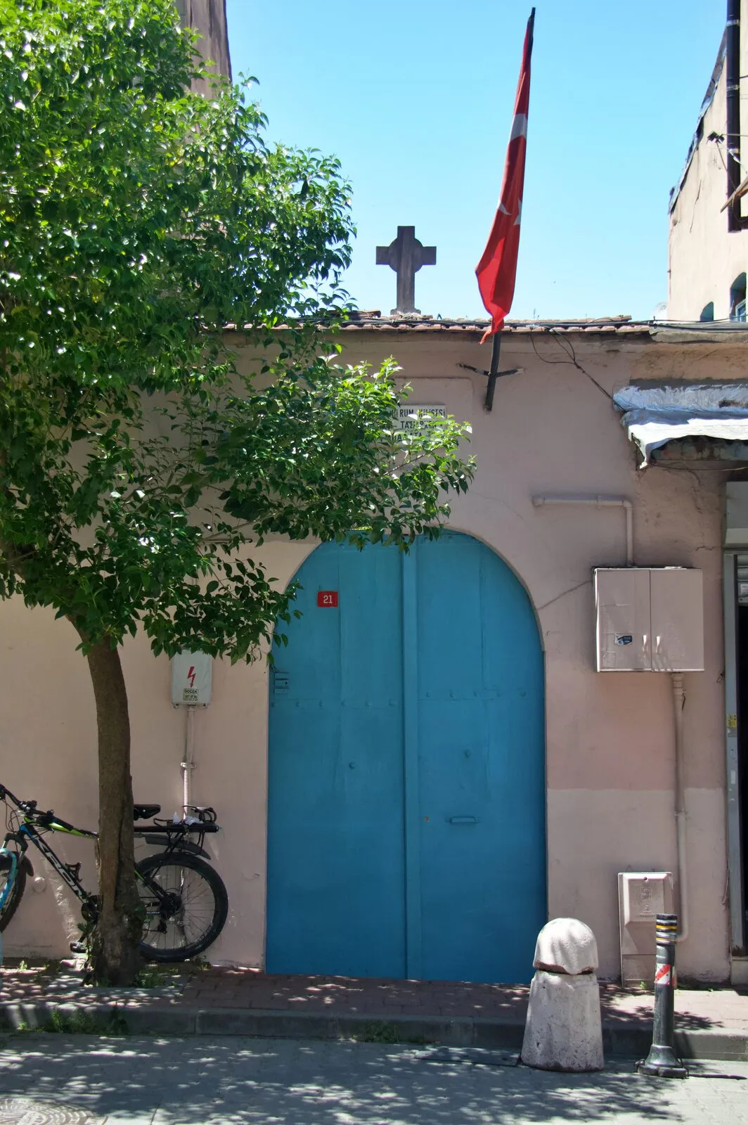 Η μπλε τοξωτή πύλη του περιβόλου του ναού στον ροζ τοίχο προς τον δρόμο, με σταυρό στη στέγη