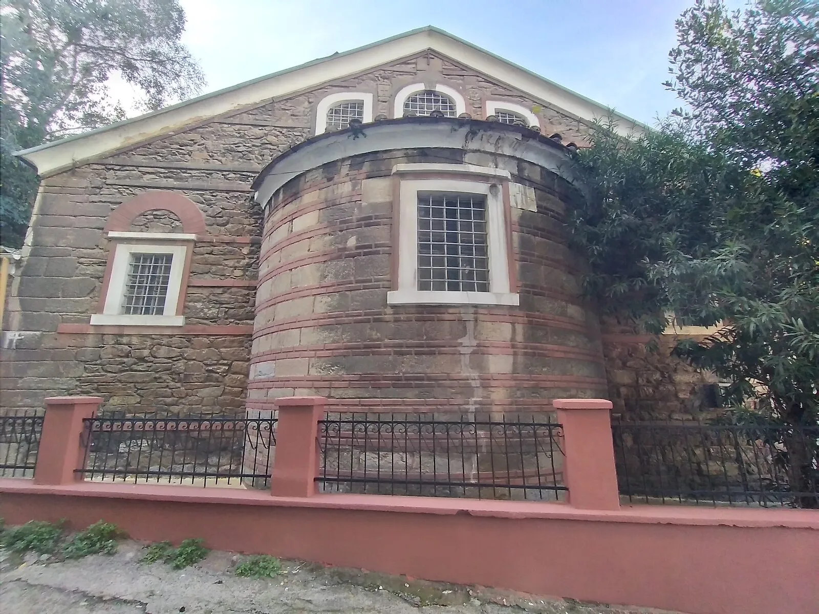 Η πέτρινη ανατολική πλευρά του ναού με την ημικυκλική αψίδα, πίσω από χαμηλό φράχτη