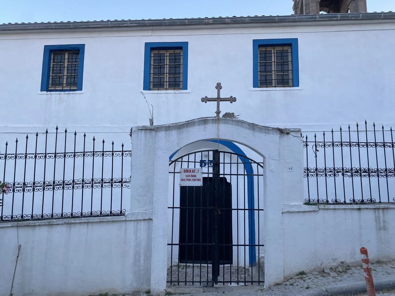 Ο λευκός ναός με τα μπλε κουφώματα πίσω από τον τοίχο της αυλής, με σταυρό πάνω από την πύλη