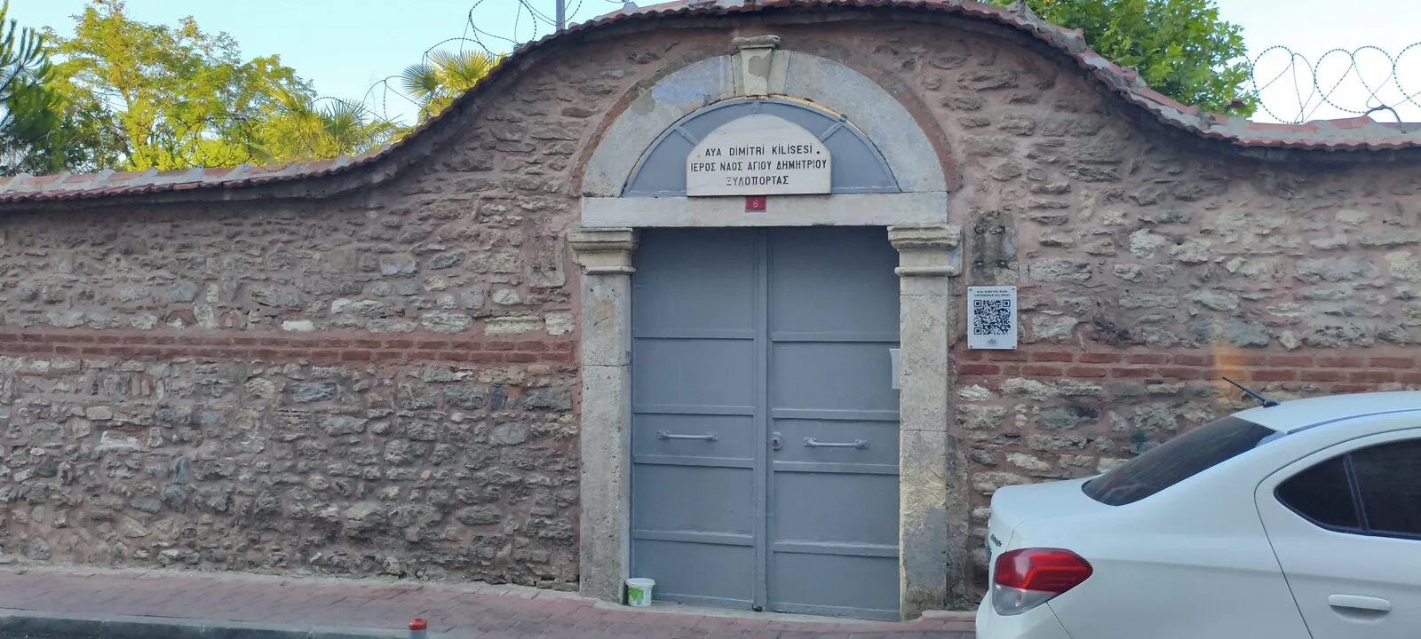 Ο πέτρινος περίβολος και η γκρίζα πύλη του ναού του Αγίου Δημητρίου, με δίγλωσση πινακίδα